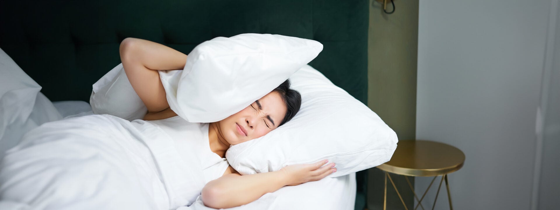 Insomnia. Asian girl shuts her ears with pillow as partner snoring. Woman lying in bed, bothered by noise at night, cannot sleep